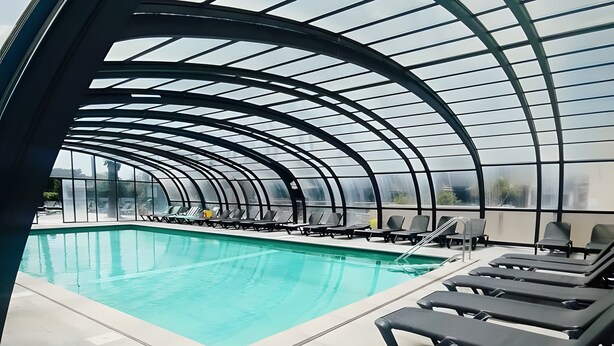 Una piscina interior moderna con techo de vidrio curvo, sillas de descanso y vista despejada del cielo.