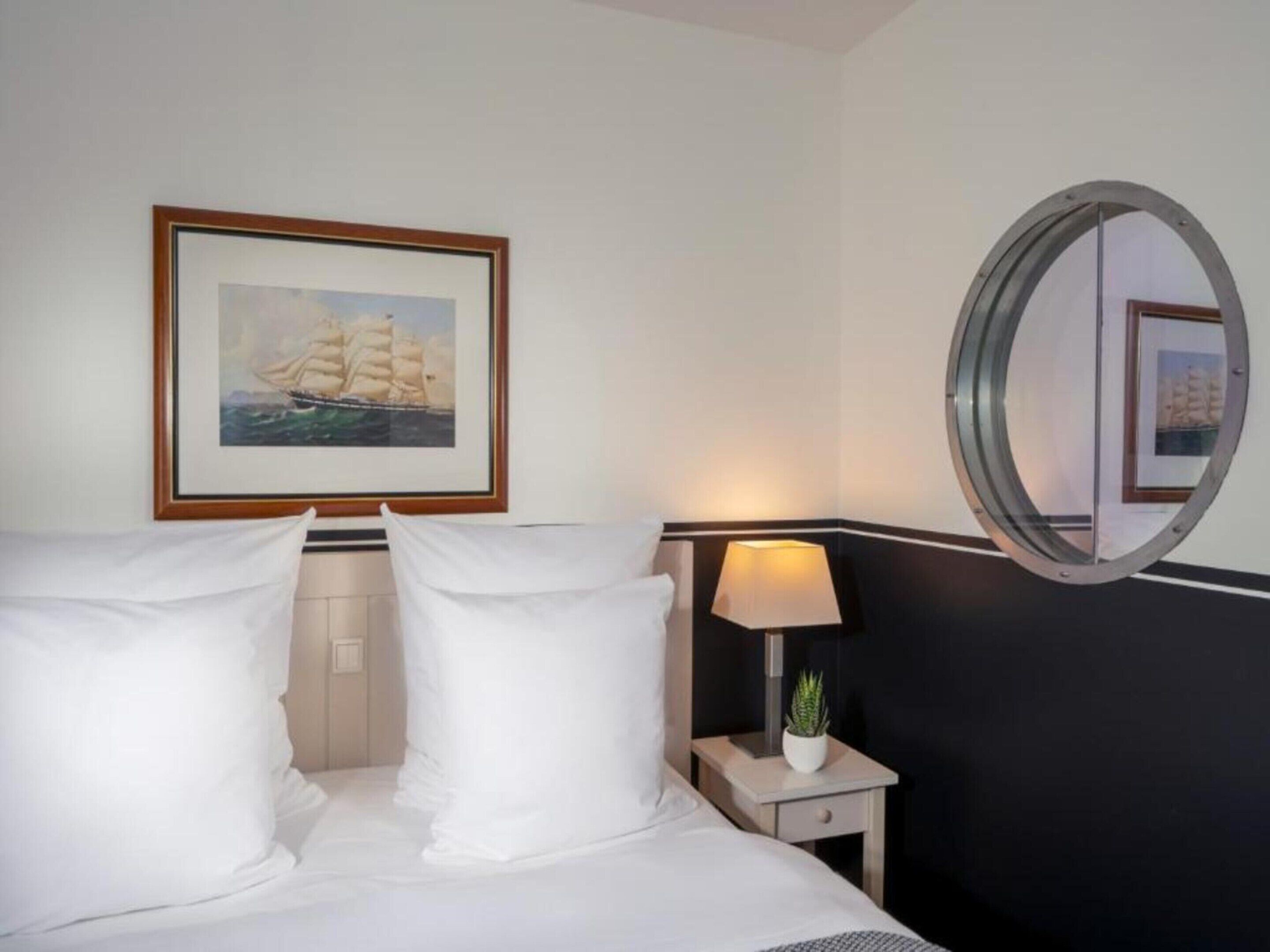 A hotel room with a bed, two pillows, a nightstand with a lamp, a small plant, and a framed picture of a sailboat on the wall.