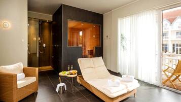 Una habitación de hotel moderna con una cama grande, un área de descanso con una silla de mimbre, una mesita con fruta y copas de vino, y un balcón con vistas a edificios.