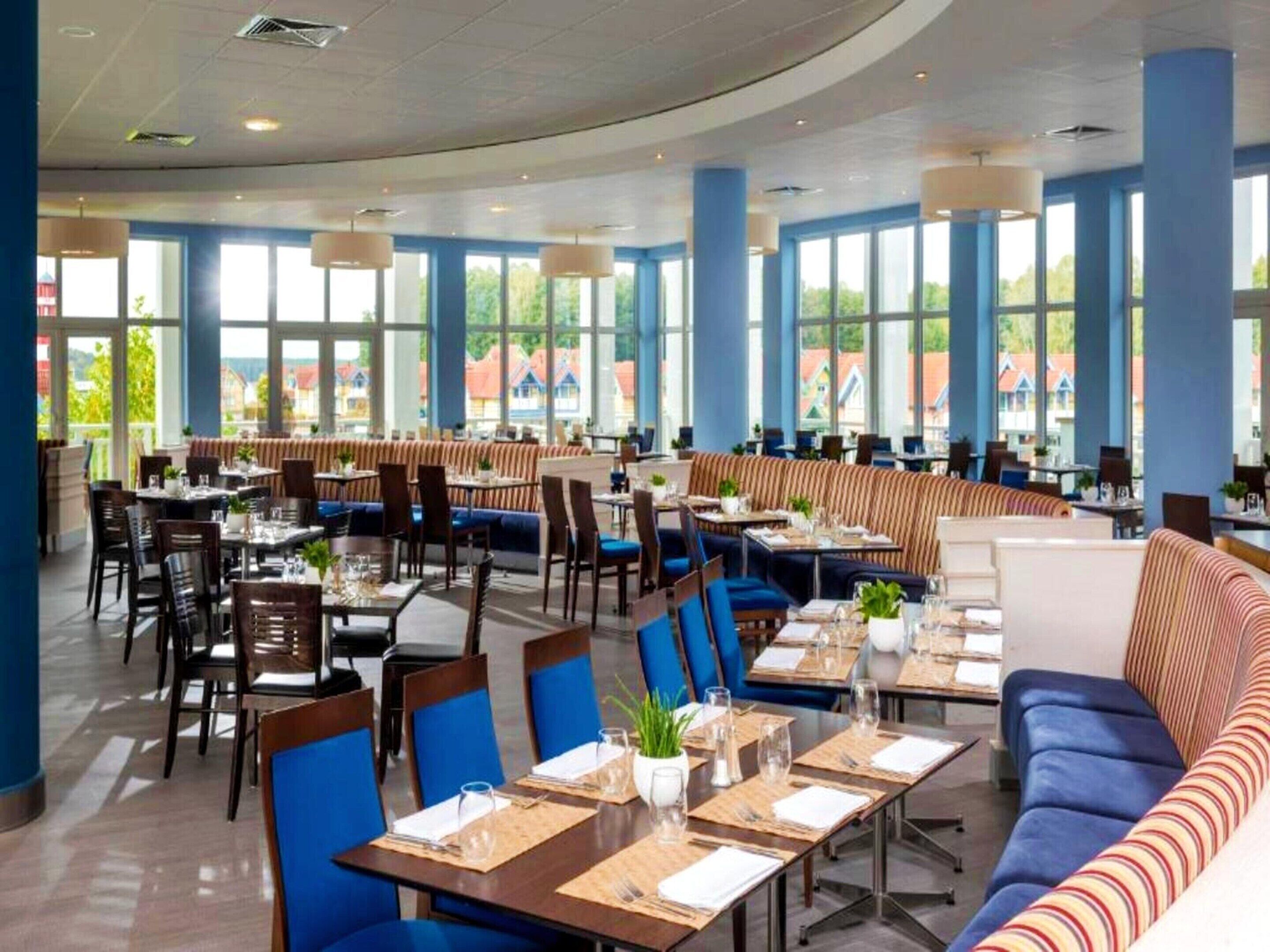 Un comedor amplio con varias mesas y sillas, ventanas grandes que ofrecen vistas al exterior y una variedad de opciones de asientos, incluyendo cabinas y una mesa larga comunitaria.