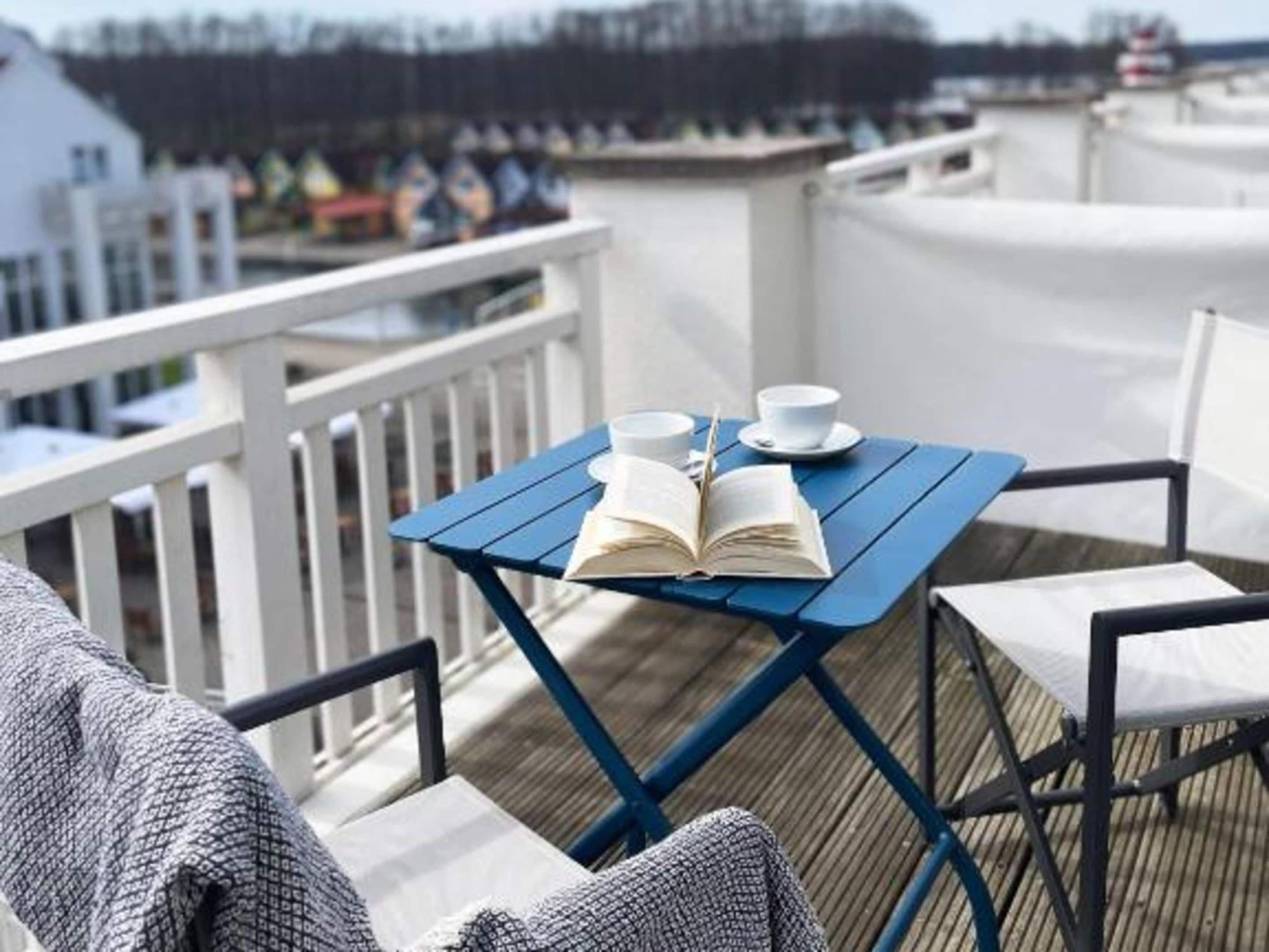 A blue table with a book and two cups on a deck with white railings.