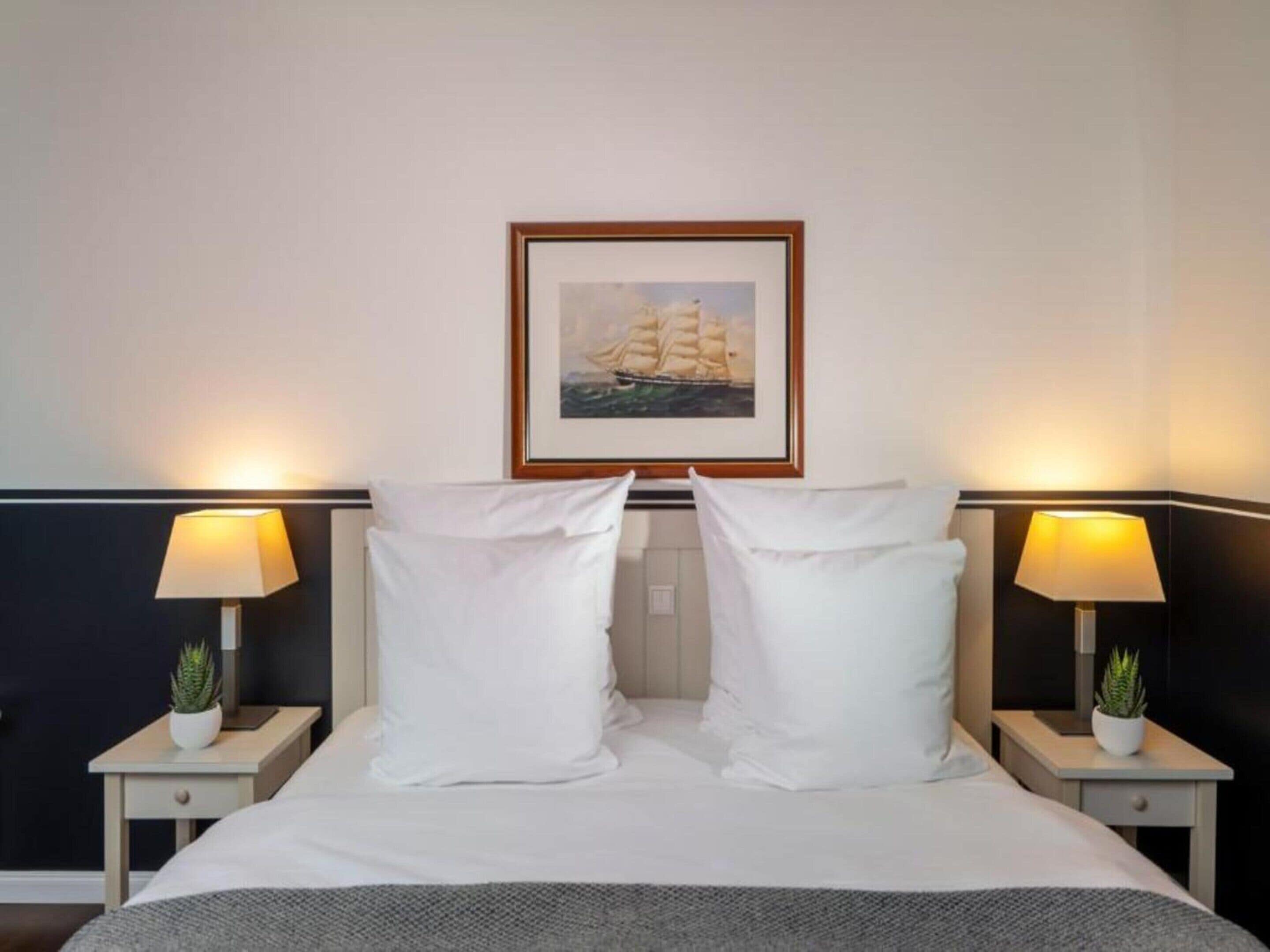 A neatly made bed with white linens, two bedside lamps, and a framed picture of a sailboat on the wall.