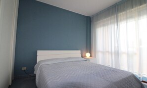 A bedroom with a bed, a nightstand, and a window with curtains. - Modern Flat With Pool View (Lignano Sabbiadoro)