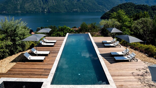 Una piscina infinita rectangular con sillones y sombrillas, rodeada de terrazas de madera y abundante vegetación, con vistas a un lago y montañas.