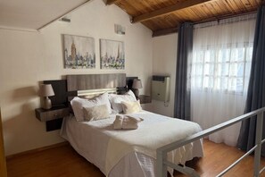 A bedroom with a wooden ceiling, a bed with white bedding, two wall-mounted paintings, a bedside table with a lamp, and a window with curtains. - Casita Temporary Rental Luz (In the center) (Río Gallegos)