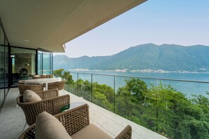 Ein Balkon mit Korbmöbeln und Glasgeländer mit Blick auf eine Berglandschaft und ein Gewässer.