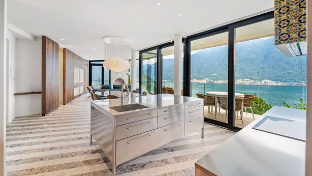 Una cocina moderna con una isla grande, un comedor con sillas de mimbre y vistas a un cuerpo de agua y montañas.