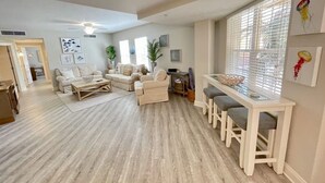 Un salon spacieux doté d’un plancher en bois, d’un ensemble de canapés, d’une table à manger avec des bancs et d’une grande fenêtre avec des stores.