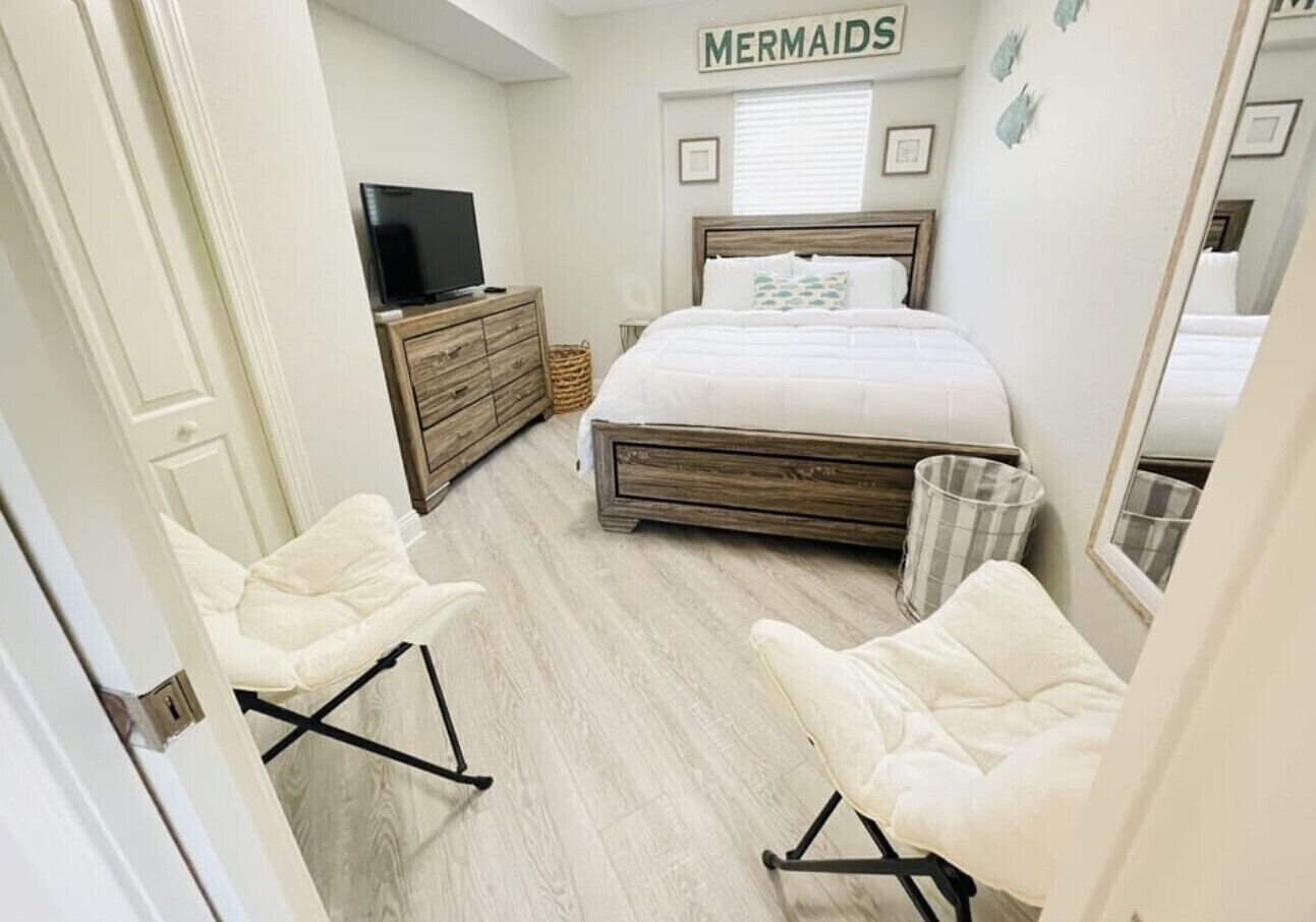 A bedroom with a bed, two white chairs, a TV, a wooden dresser, and a wall sign that reads "MERMAIDS".