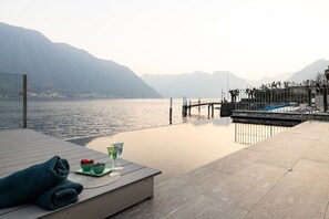 Une terrasse paisible au bord du lac, avec une plateforme en bois, une table en verre sur laquelle se trouve un saladier rempli de fruits et un verre, ainsi qu’une serviette enroulée.