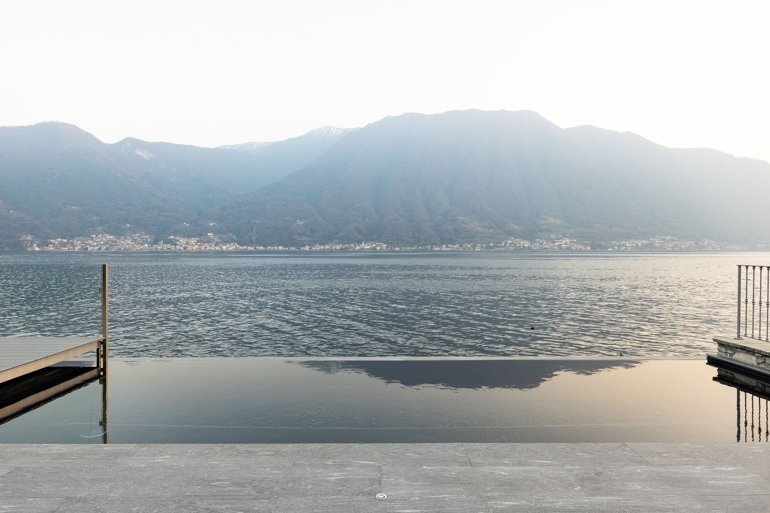 A waterfront view with a dock extending into calm water, surrounded by mountains.