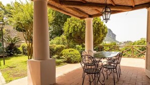 Eine Terrasse im Freien mit Tisch und Stühlen, umgeben von Grünpflanzen und Blick auf einen Berg.