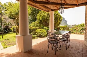 Eine Terrasse im Freien mit Tisch und Stühlen, umgeben von Grünpflanzen und Blick auf einen Berg.