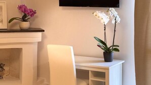 Une pièce avec un bureau blanc, une chaise, un téléviseur à écran plat fixé au mur et deux plantes en pot ornées de fleurs.
