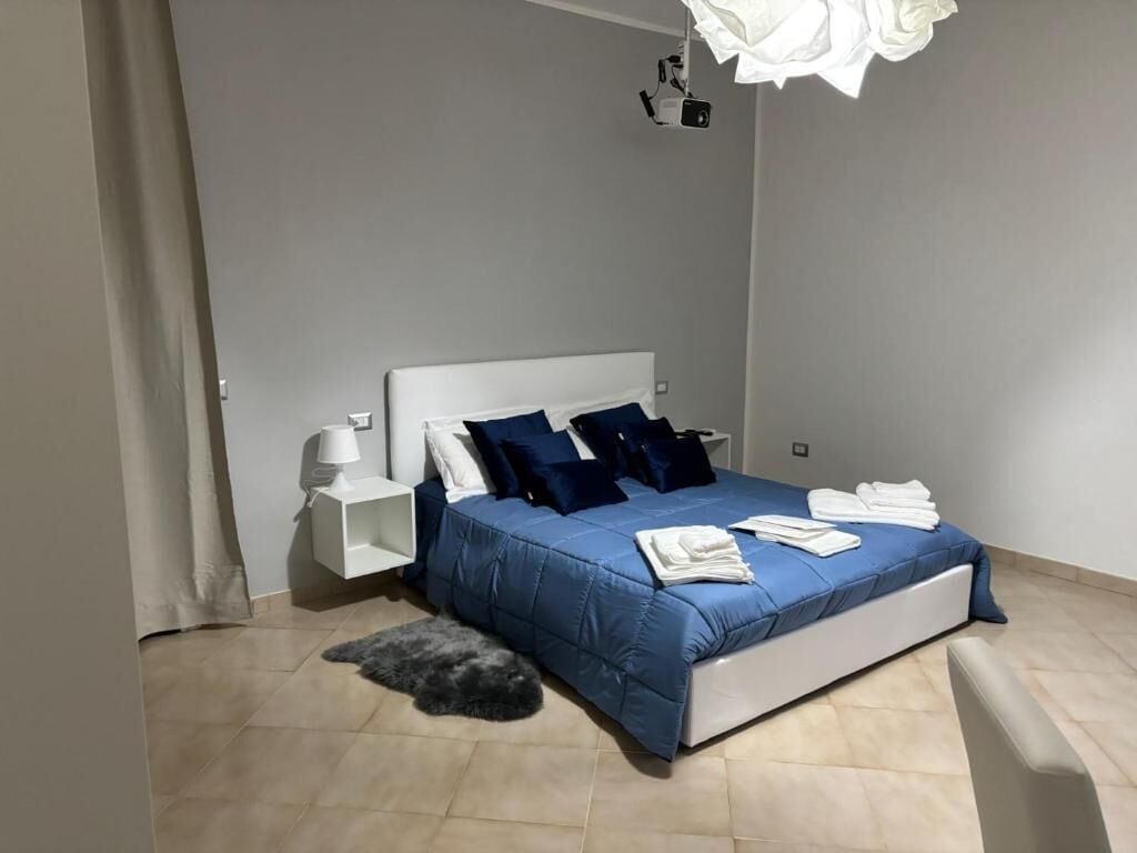 A bedroom with a bed, white nightstands, a lamp, and a ceiling-mounted projector.
