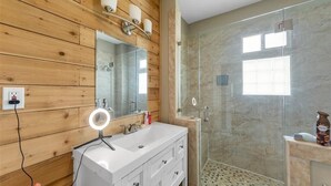 Un baño con pared de madera, tocador blanco, espejo con iluminación, cabina de ducha con mampara de vidrio y una ventana pequeña.