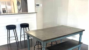 Un comedor con una mesa de madera y banco, dos taburetes negros, y una cocina al fondo.
