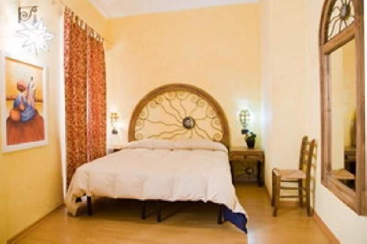 A bedroom with a bed, a chair, a nightstand, a mirror, and a wall with a decorative headboard.