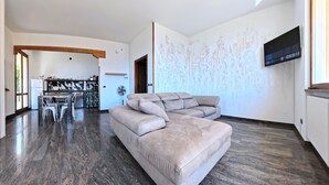 A modern living room with a large L-shaped sofa, a dining area with a table and chairs, and a kitchen visible through an open doorway. - Villa Arianna (Gardola)
