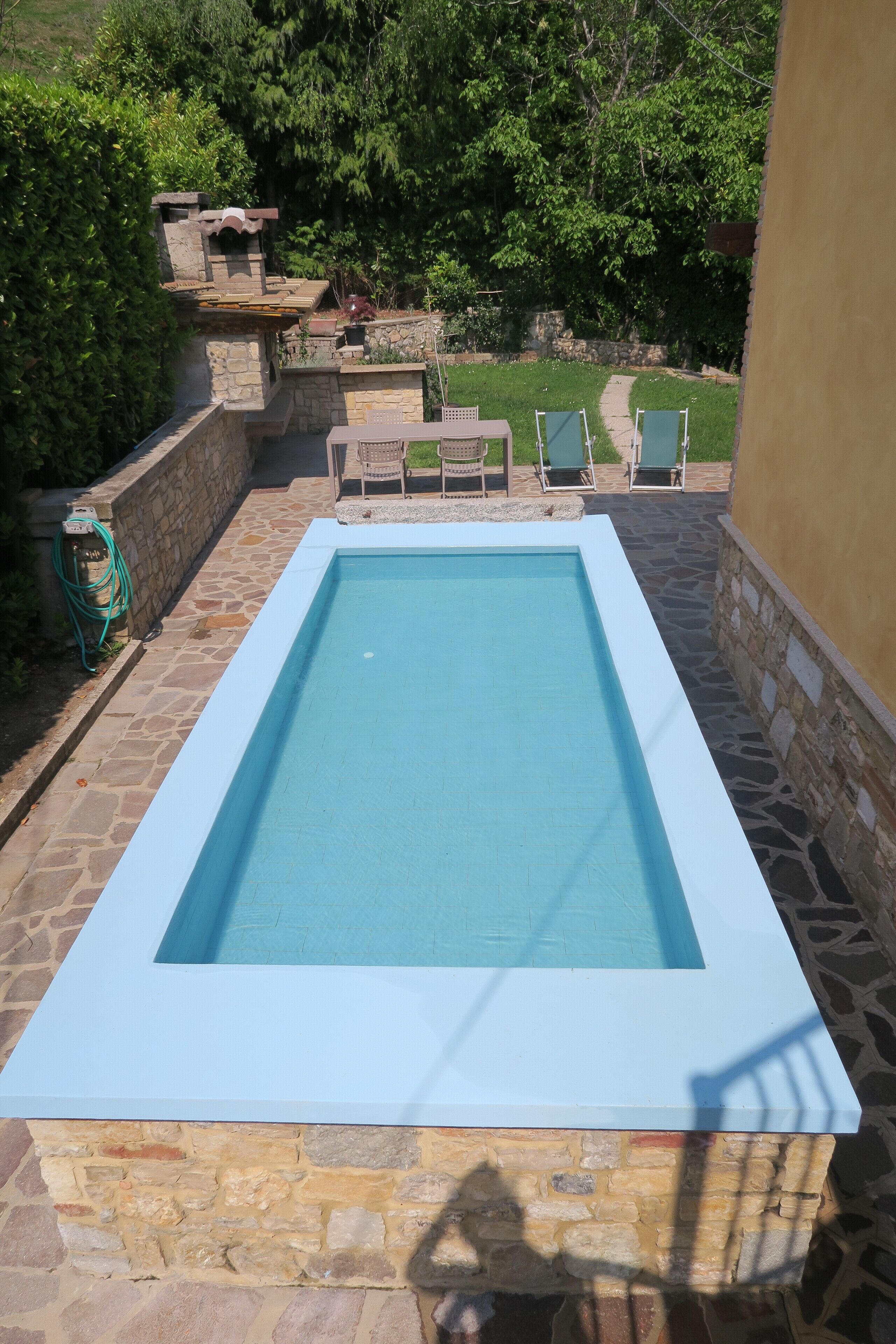Une piscine rectangulaire bordée de pierres et de béton, entourée d’une terrasse en pierre et d’un petit espace de détente avec des chaises.