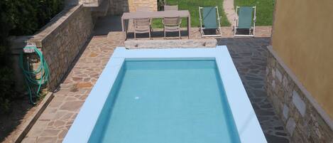 Une piscine rectangulaire bordée de pierres et de béton, entourée d’une terrasse en pierre et d’un petit espace de détente avec des chaises.