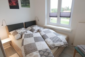 Ein Doppelbett mit Patchwork-Quilt, ein Nachttisch, eine Lampe, ein Fenster mit Jalousien und ein Heizkörper.