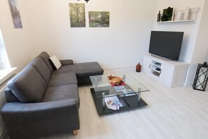Ein modernes Wohnzimmer mit einem grauen Ecksofa, einem Glastisch mit Zeitschriften, einem Flachbildfernseher auf einem weißen Schrank und gerahmten Naturfotos an der Wand.