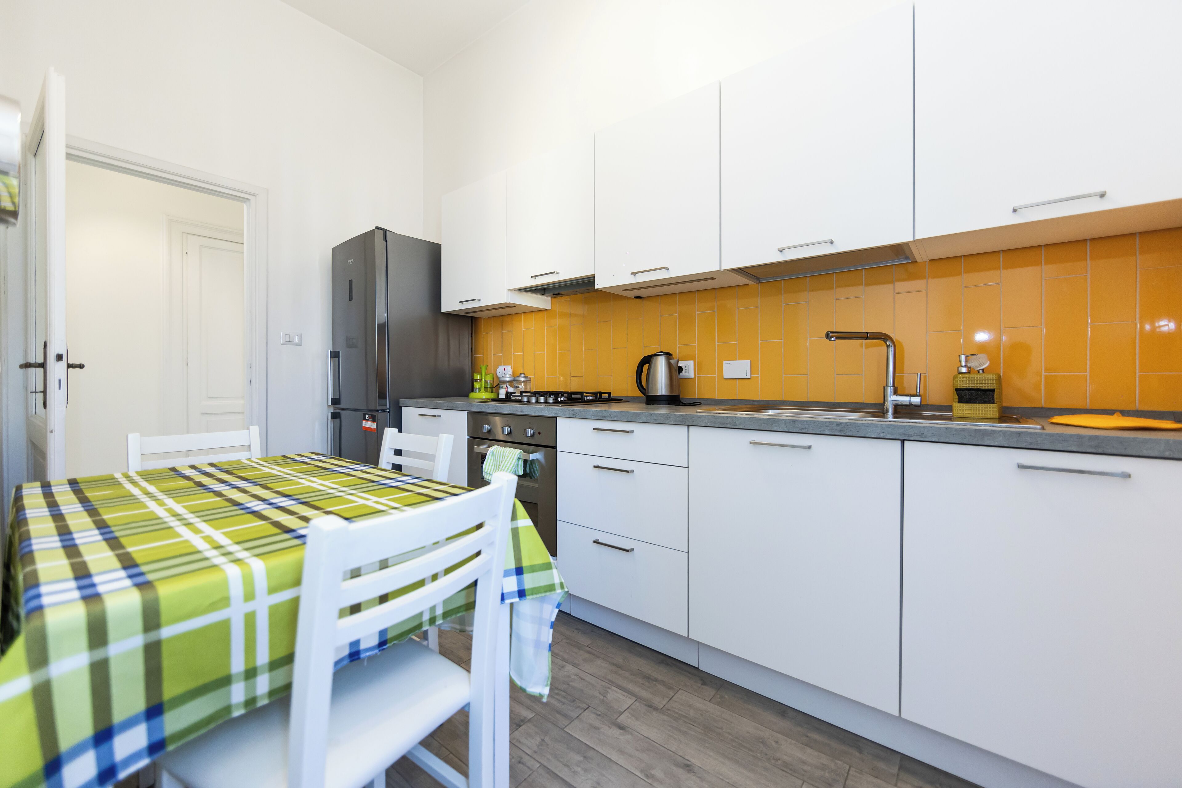 Een kleine keuken met witte kastjes, een roestvrijstalen koelkast en een backsplash met gele tegels.