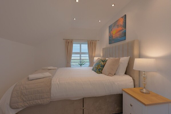 A bedroom with a bed, a nightstand, a lamp, a painting, and a window with curtains.