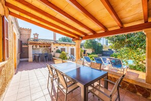 Un patio exterior con una pérgola de madera, una mesa de comedor con sillas, una piscina y vistas a un edificio tradicional con tejado de tejas.