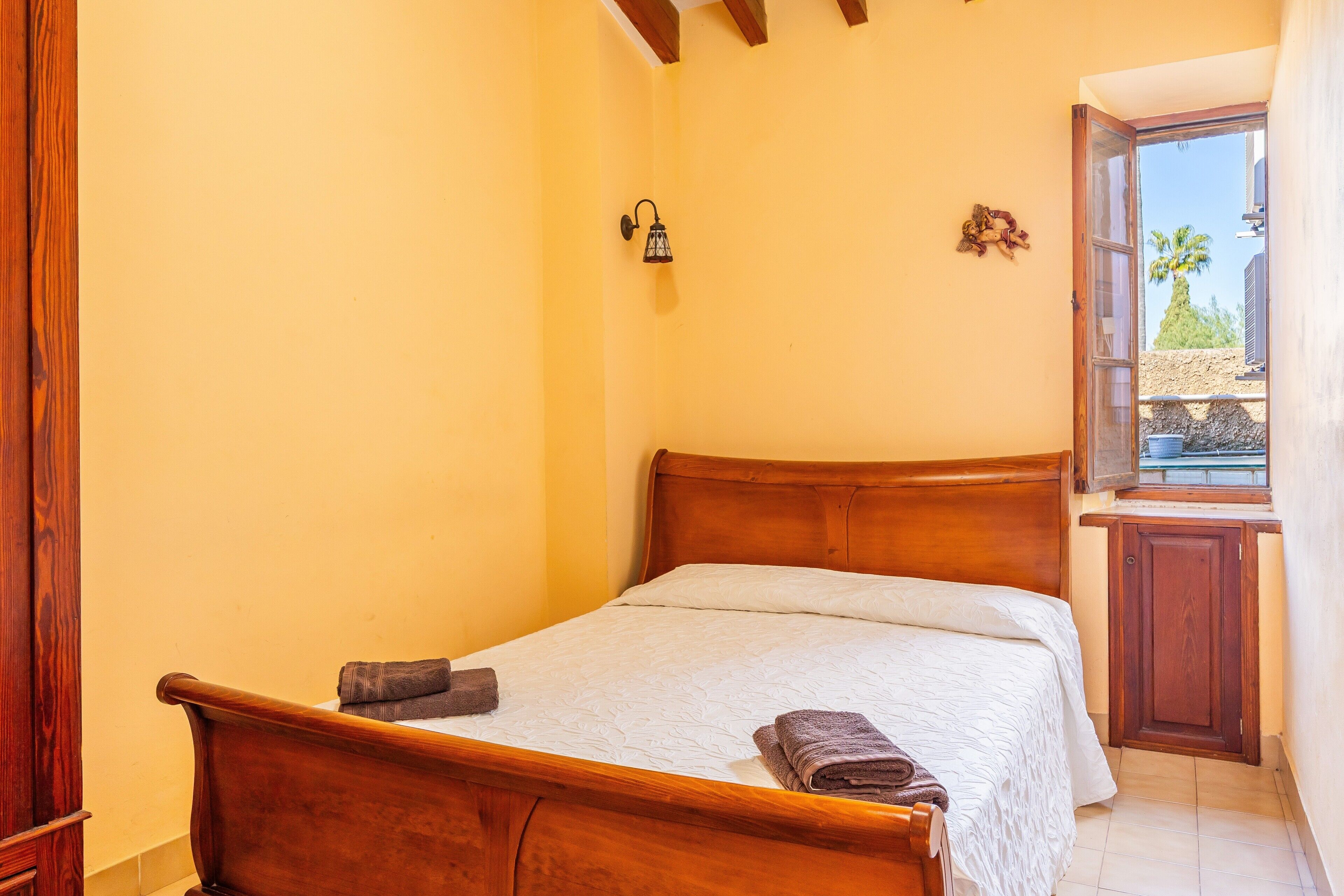 Une chambre avec un lit en bois, une table de chevet, une lampe fixée au mur et une fenêtre donnant sur des palmiers.