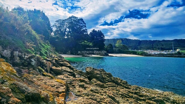 Paisatge costaner amb terreny rocos, aigües blaves cristal·línes i una platja a la distància.