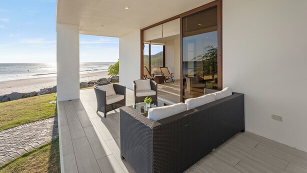 A balcony with wicker furniture, a glass door leading to a room, and a view of the beach. - Hacienda Iguana Beachfront 3BR · Surf & Sunset (Tola)