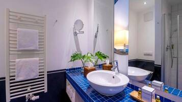 A bathroom with a white sink, two mirrors, a towel rack with white towels, a potted plant, and a shower area.