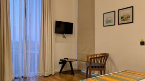 A hotel room with a wooden floor, a bed with a striped bedspread, a chair, a small table with a lamp, a TV on the wall, and a window with sheer curtains. - Private bedroom with ensuite bathroom in Colombo 5 (Colombo)