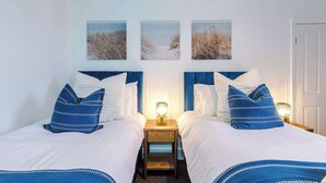 Quarto duplo com duas camas, cada uma com edredom branco e roupa de cama azul. Três quadros pendurados na parede retratam cenas de praia.