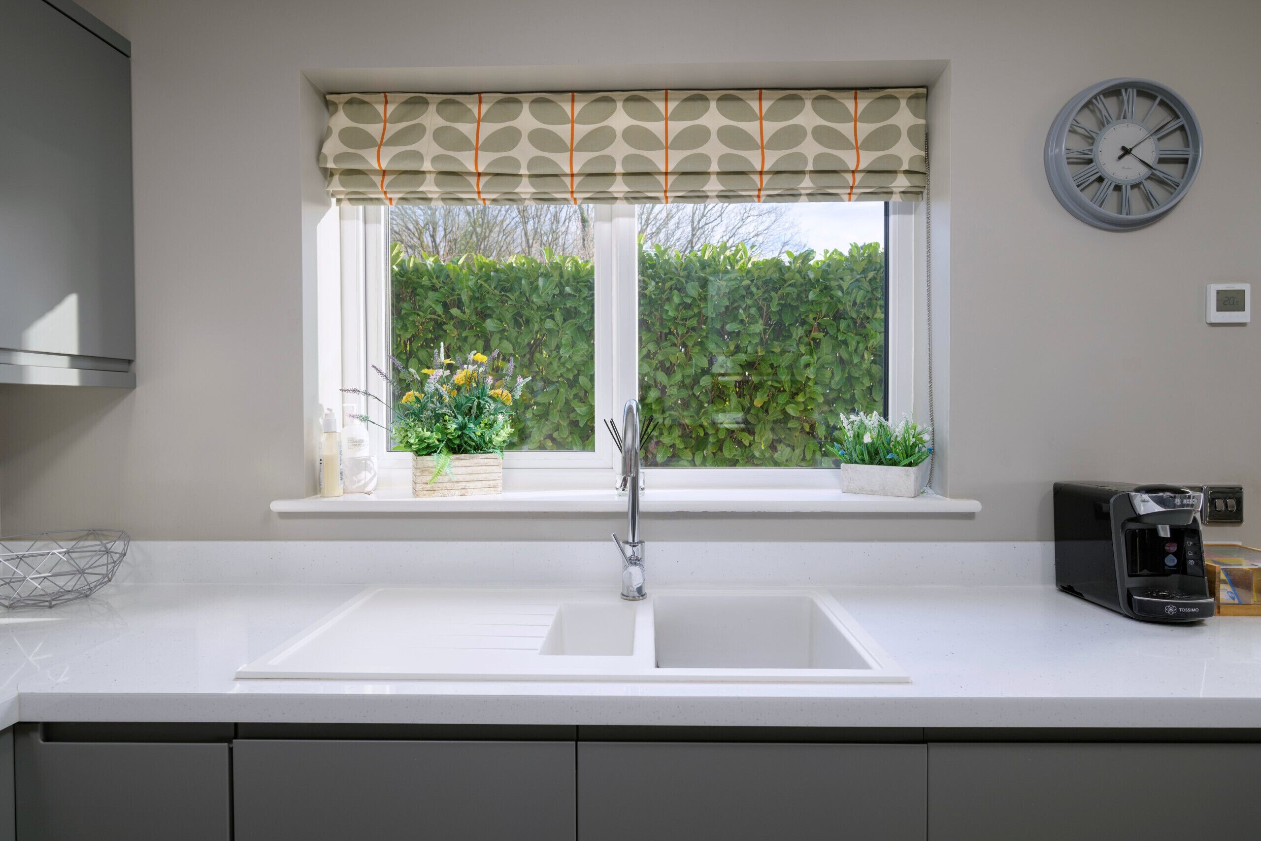 A kitchen with a double sink, a window with a leaf-patterned valance, a wall-mounted clock, and a coffee machine.