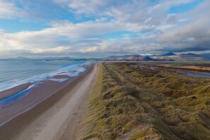Eine Küstenlandschaft mit Strand, grasbewachsenem Gelände und Bergen in der Ferne, bewölkter Himmel.