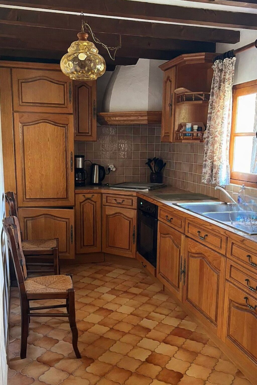 Een keuken met houten kastjes, een betegelde spatwand, een koffiezetapparaat en een raam met gordijnen.
