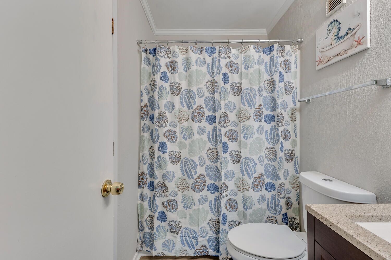 Une salle de bain avec un rideau de douche orné d’un motif de coquillages, des toilettes blanches et un plan de toilette beige.