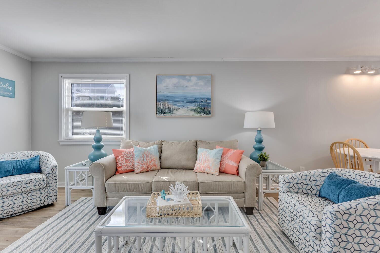 A living room with a beige sofa, patterned armchairs, a glass coffee table, and a painting of a beach scene.