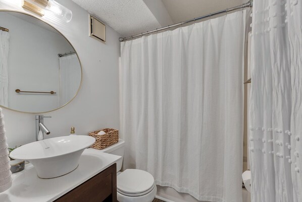 Un baño con un lavabo redondo, inodoro, espejo y cortina de ducha.