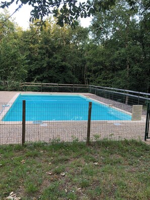 Une piscine rectangulaire entourée d’une clôture en fil de fer, avec une pelouse au premier plan et des arbres denses en arrière-plan.