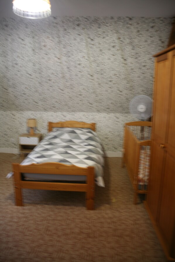 Une chambre à coucher comprenant un lit, une armoire en bois, une table de chevet avec une lampe et un ventilateur fixé au mur.