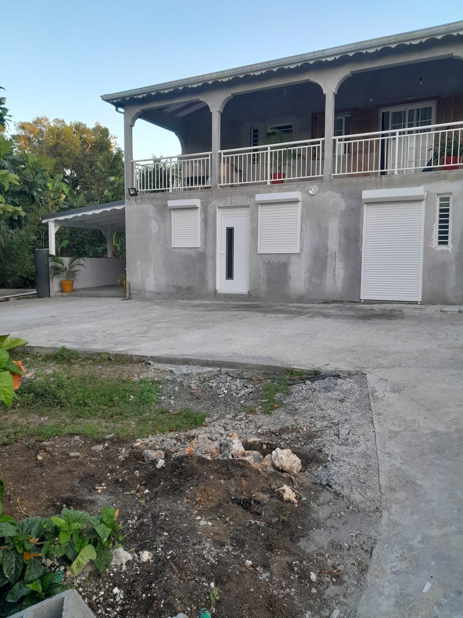 Un edificio a due piani con un balcone coperto, un'autorimessa e un piccolo giardino con piante.