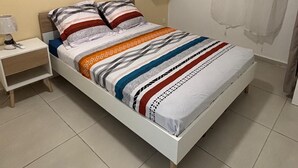 Yhden hengen huone, jossa on valkoinen sänky, raidallinen vuodevaatteisto, yöpöytä lampun kanssa, seinälle kiinnitetty peili ja ilmastointilaite.