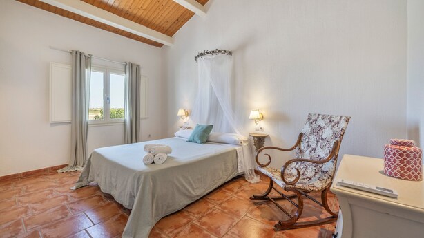 Un dormitorio con cama, una silla, una mesita de noche, un ventanal con cortinas y vistas al exterior.