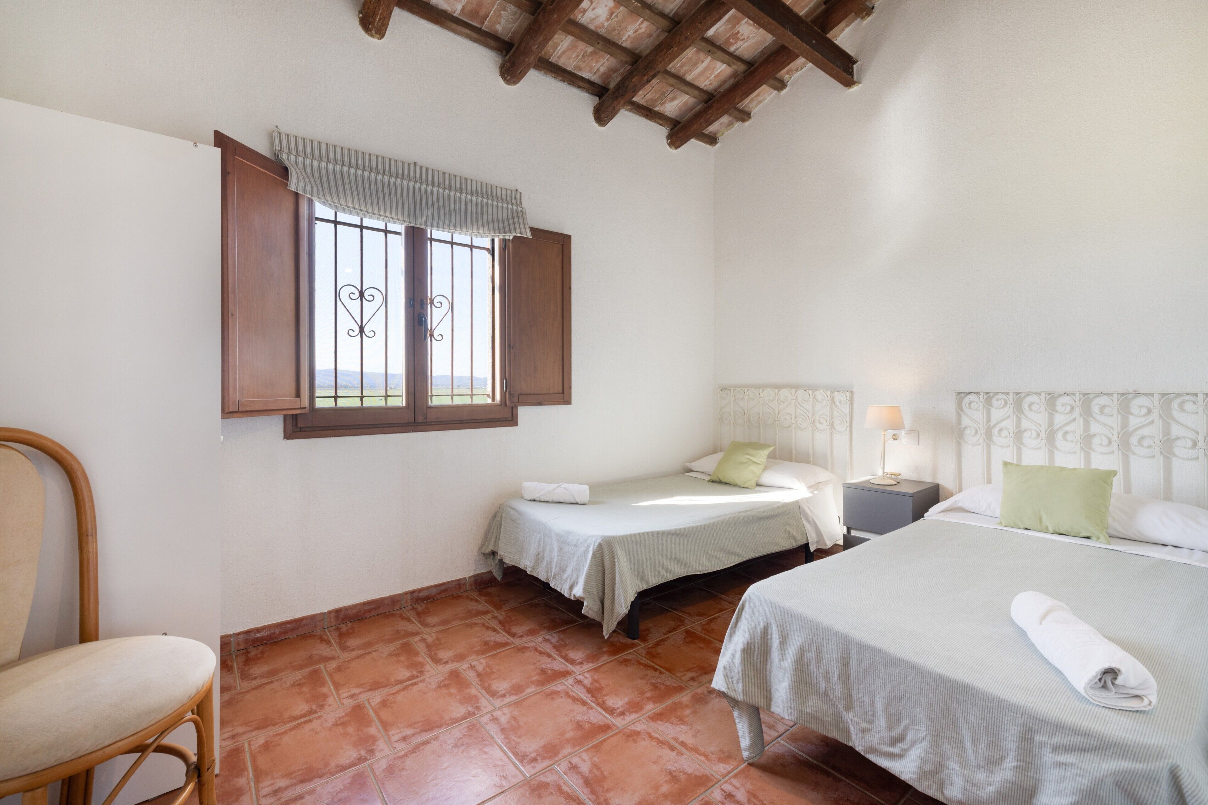 A bedroom with two beds, a chair, a window with shutters, and a ceiling with exposed beams.