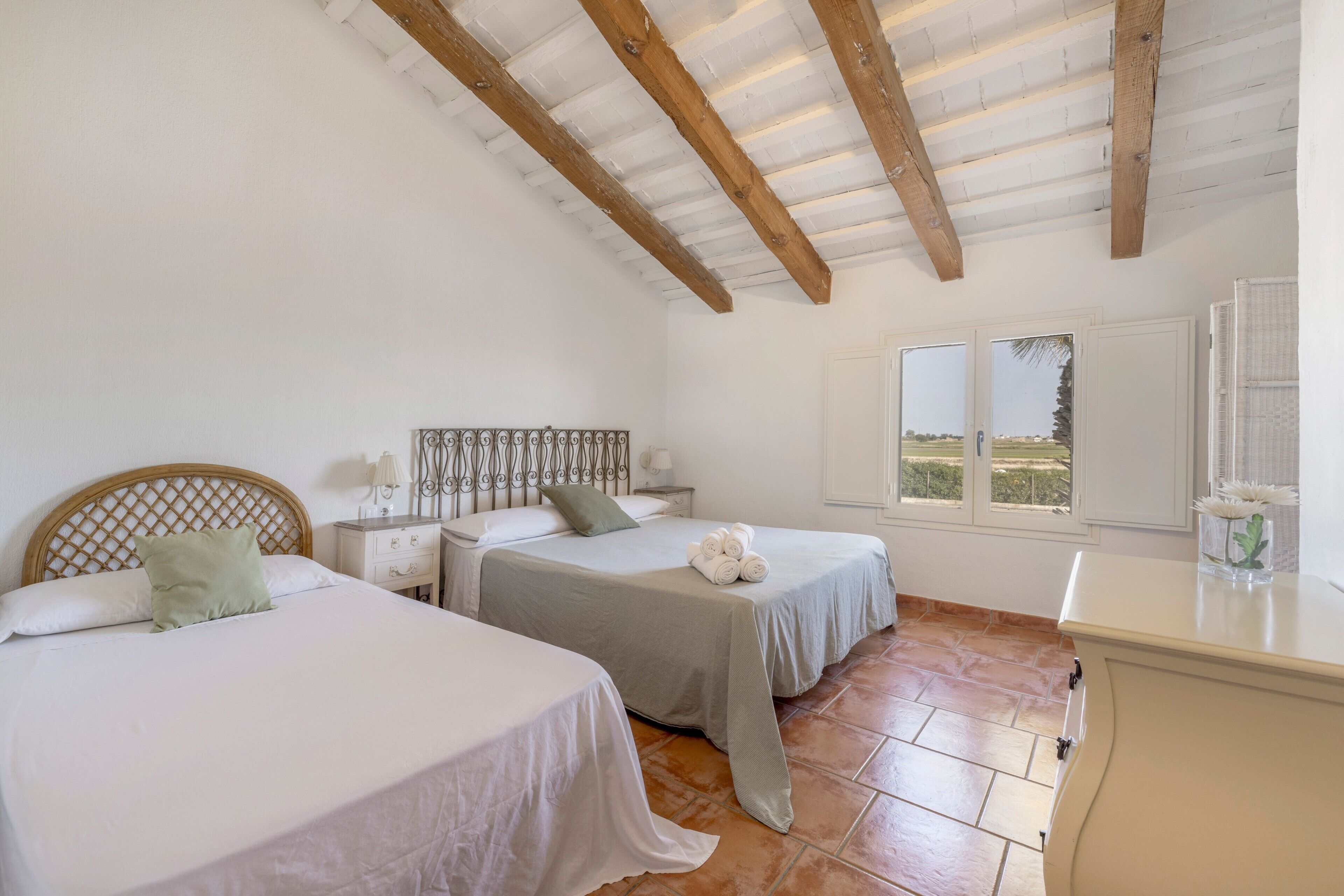 A bedroom with two beds, a wooden ceiling, a window with a view, and a bedside table with a lamp.