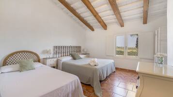A bedroom with two beds, a wooden ceiling, a window with a view, and a bedside table with a lamp.